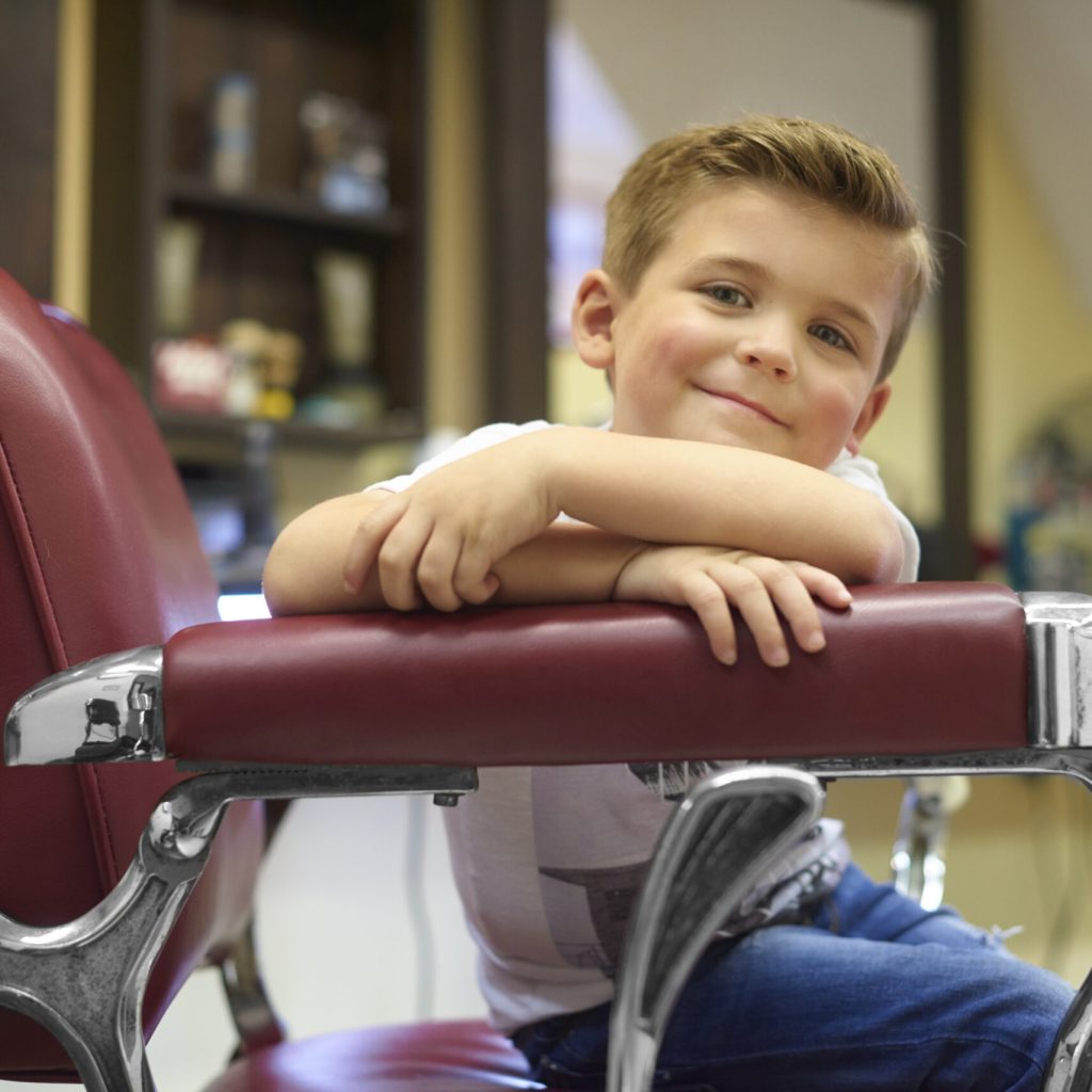 A young boy has had his hair cut.He is very happy that he got to sit in the barbers chair.He is very pleased with his new haircut.